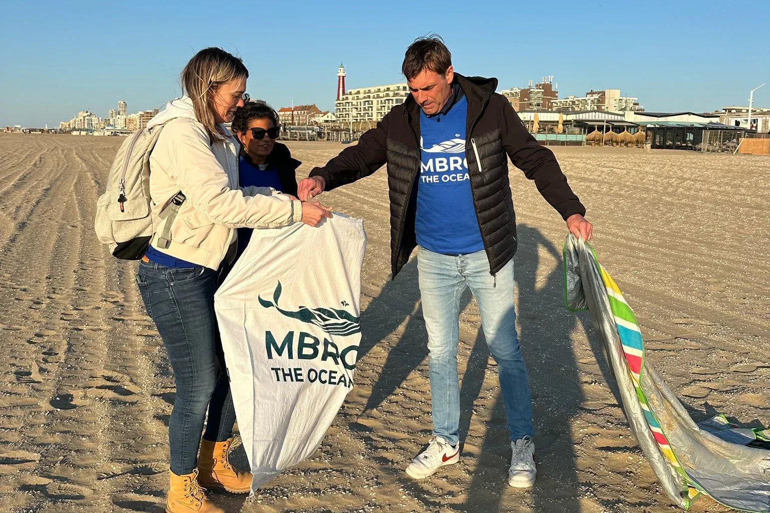 Drie mensen ruimen afval op het strand op, met stad en vuurtoren op de achtergrond.