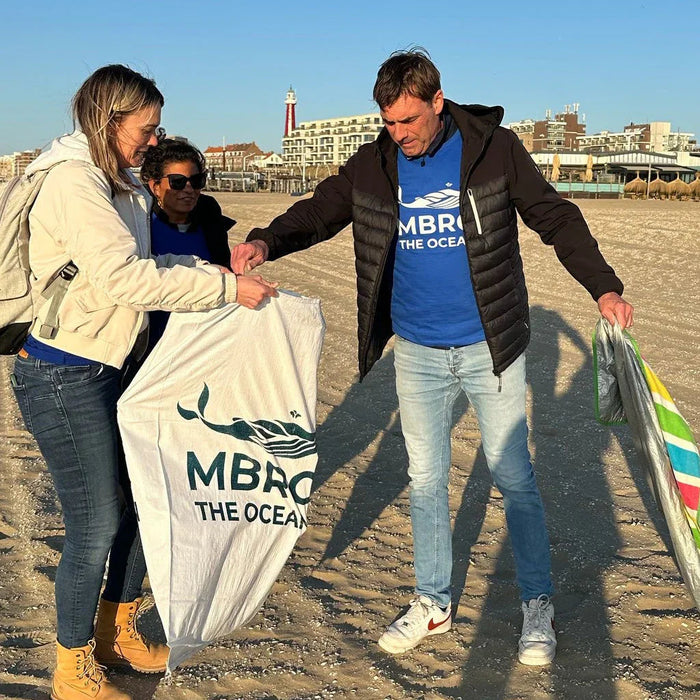 Drie mensen ruimen afval op het strand op, met stad en vuurtoren op de achtergrond.