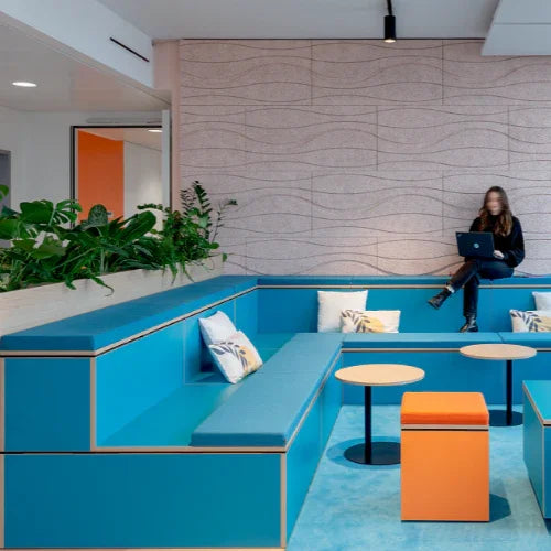 Modern kantoormeubilair met blauwe zitbanken, oranje elementen en planten in loungehoek.
