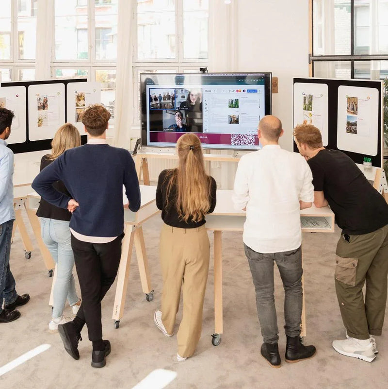 Team rond verrijdbare bureaus kijkt naar groot scherm in moderne kantoorruimte